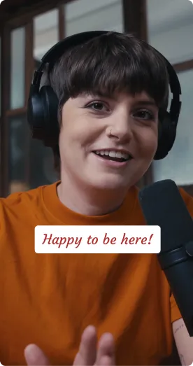 Person with short dark hair wearing headphones and an orange shirt, speaking into a microphone with a smile, with text overlay saying 'Happy to be here!'